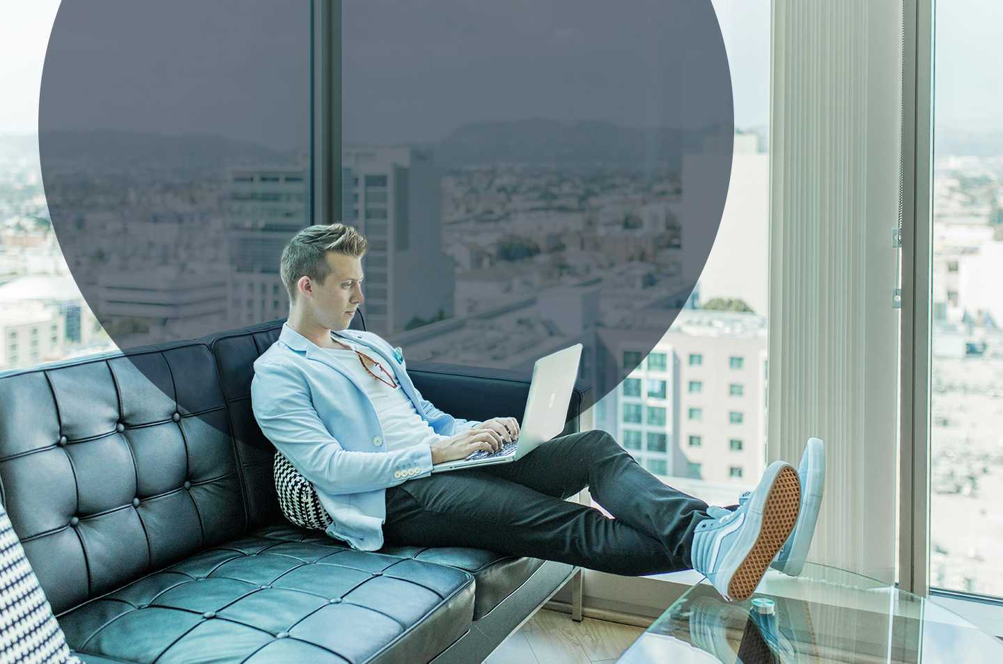 a man sitting on a couch working on his laptop with a city skyline in the background